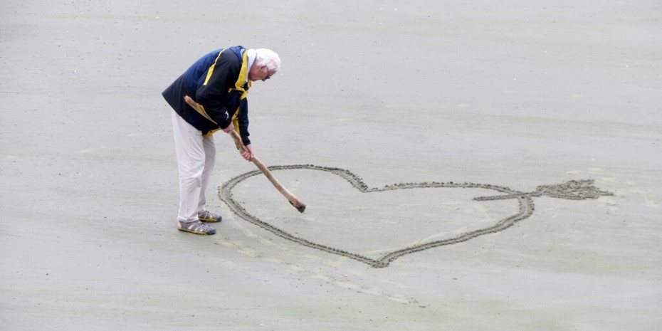 Elderly man drawing heart shape in sand at beach
