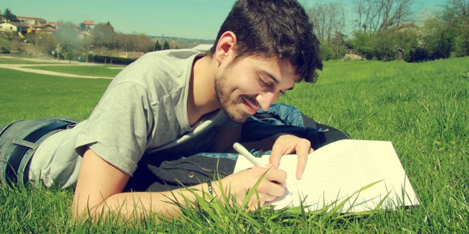 Young man with short dark hair wearing grey shirt laying on grass writing in journal