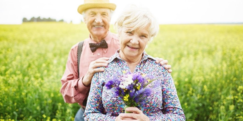 Elderley man wearing hat and elderly woman holding flowers in a yellow field