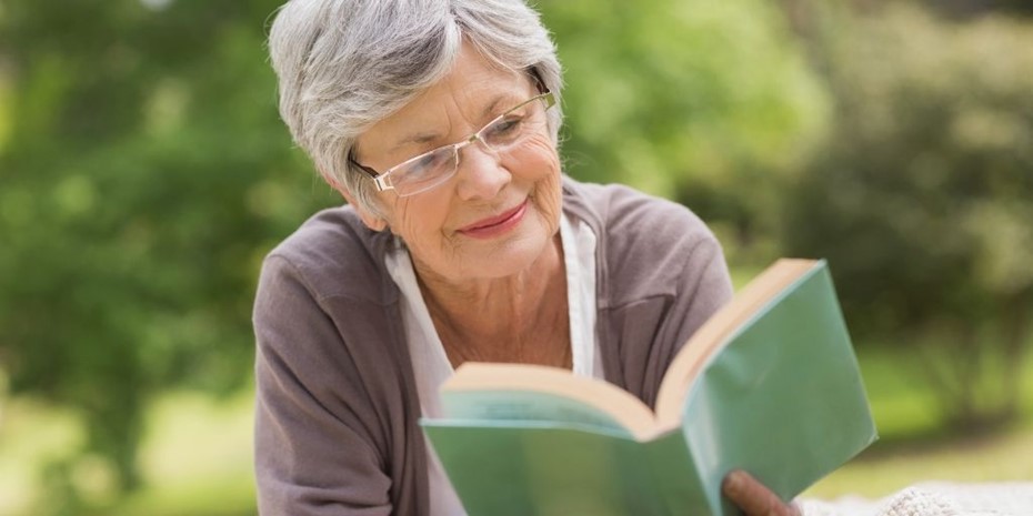 Elderly woman with grey hair and glasses reading a book with a plain green cover outside