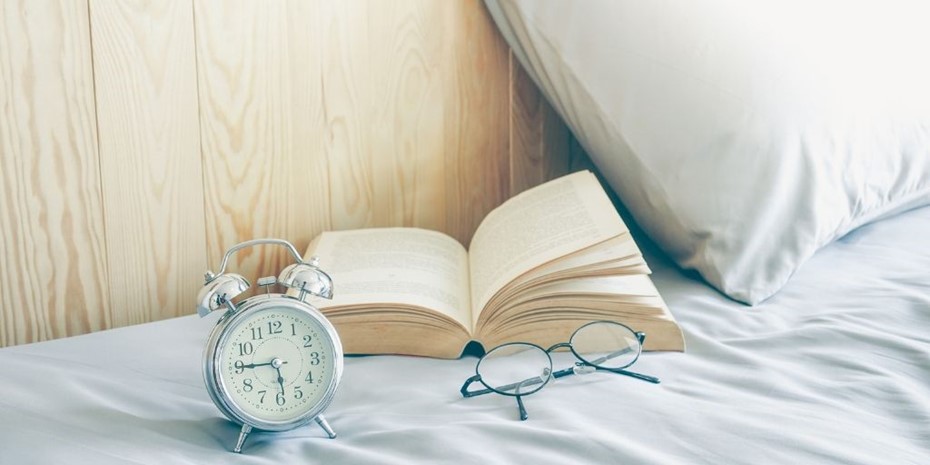 Book laying open next to pillow, reading glasses and an alarm clock
