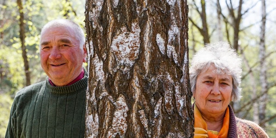 Elderly couple smiling standing either side of a tree