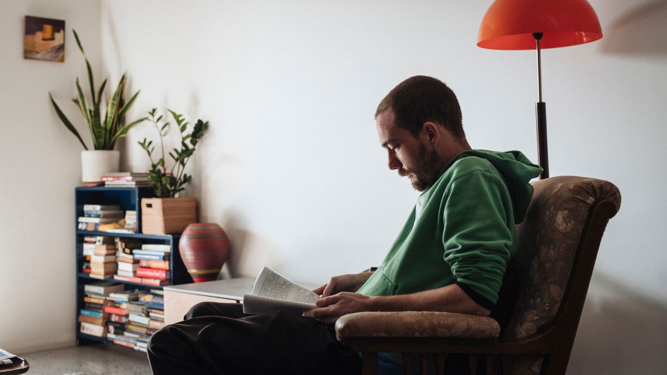 Brightwater Psychology_disability client reading book comfortably on armchair with tall orange lamp, bookshelf and indoor plants in living space